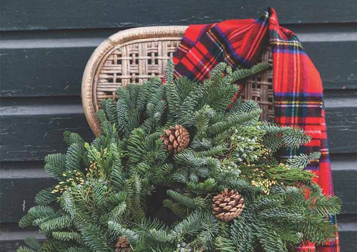 An evergreen wreath with pinecones sitting in a chair with a plaid scarf.