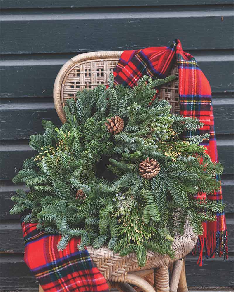 An evergreen wreath with pinecones sitting in a chair with a plaid scarf.