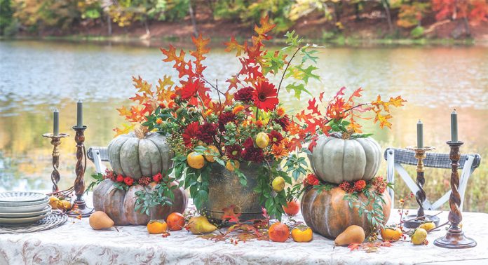 A close-up of an alfresco tablescape with stacked pumpkins, a centerpiece with fall foliage, and scattered fruit.