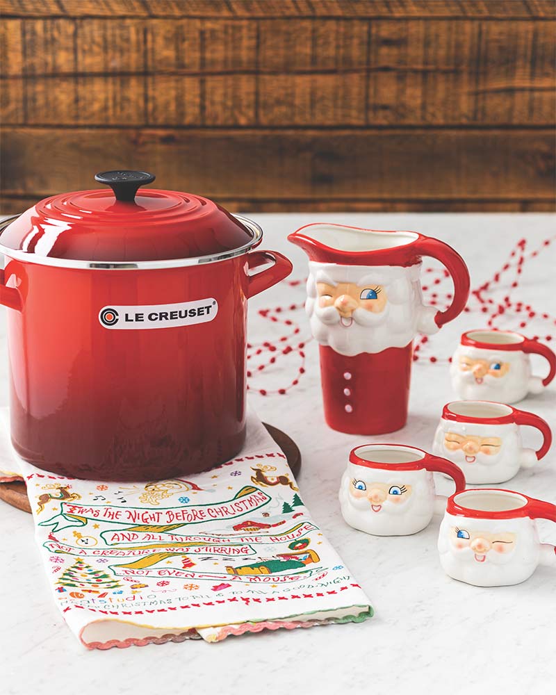 A red stockpot, a Christmas dish towel, and a Santa pitcher with matching miniature mugs. 