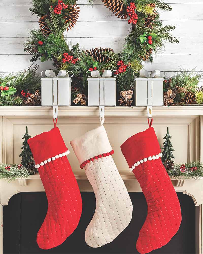 A mantel with a garland, wreath, present shaped stocking holders, and three red-and-white stockings.