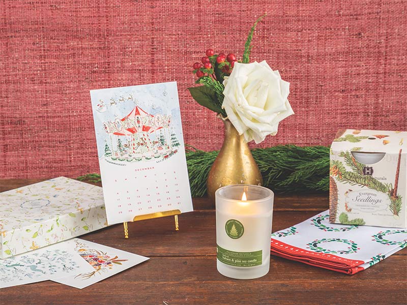 Stationery, a desk calendar, a candle, tea towel with holiday wreaths, and flowers in a gold bud vase. 