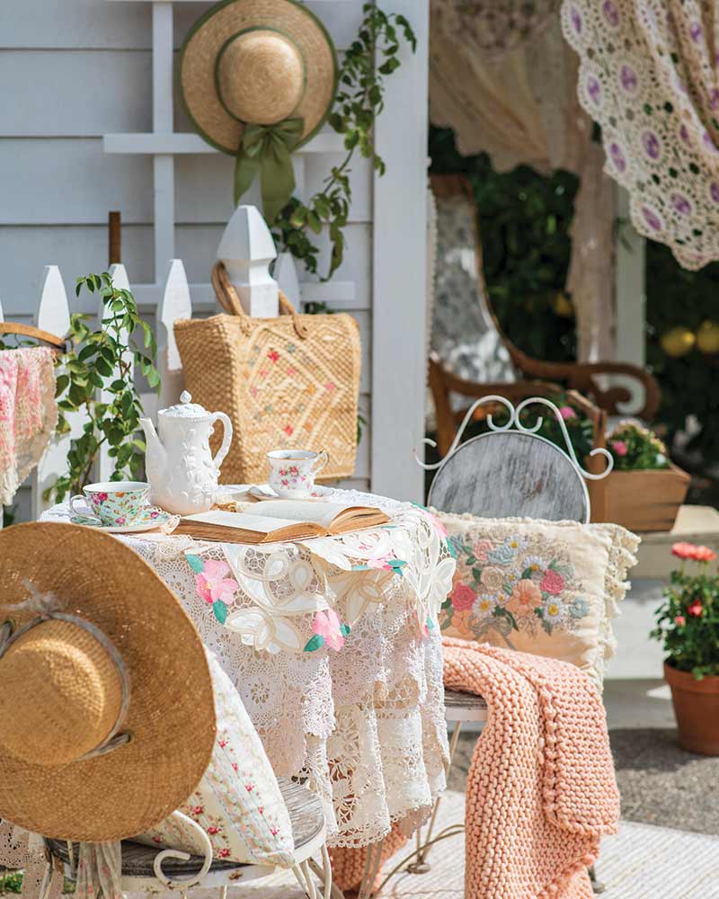 An alfresco table set for tea with pillows, a knit blanket, and sun hats. 