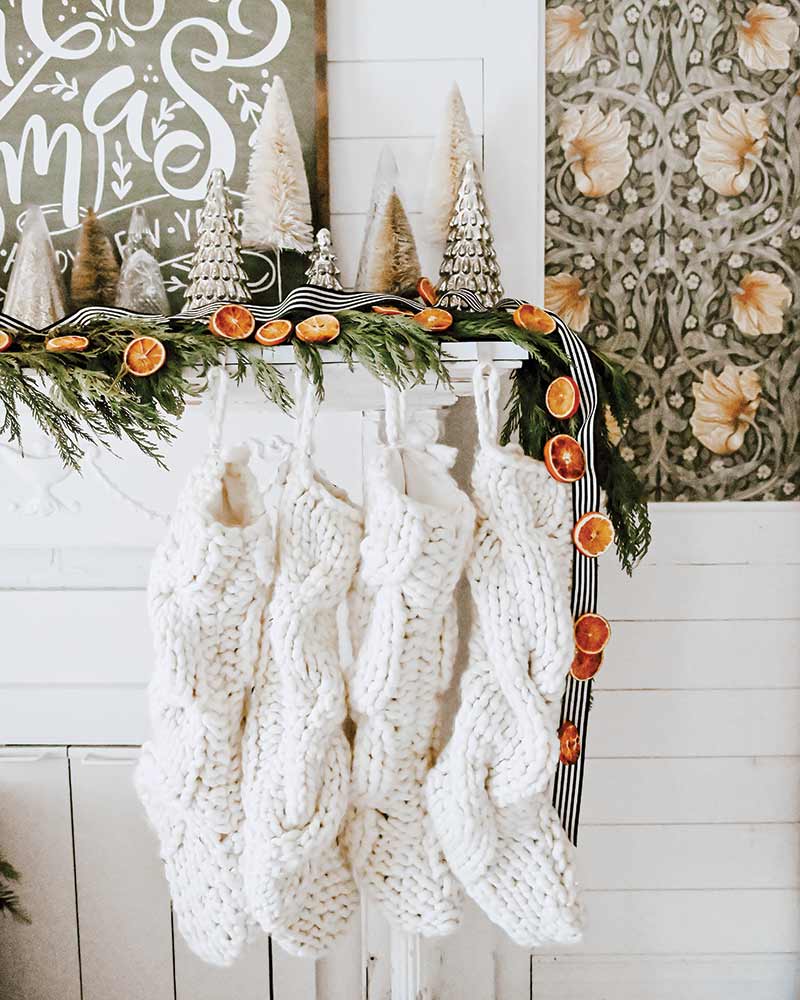 A mantle decorated with greenery, a garland of orange slices, and four white knit stockings. 
