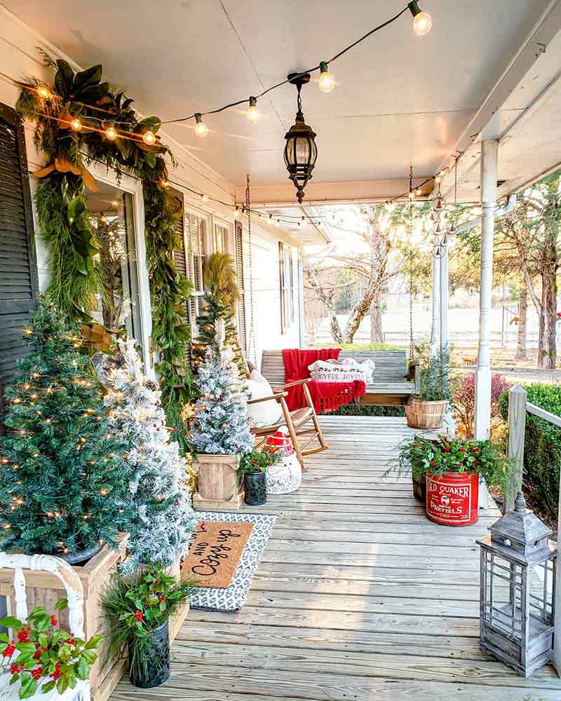 A front porch decorated with miniature trees, string lights, and festive throw pillows.