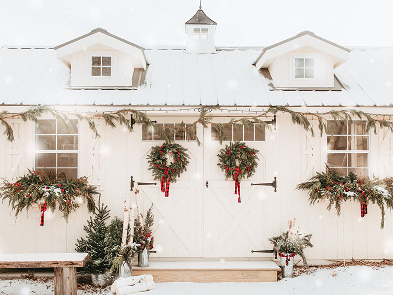 A white snow-covered barn with a pair of wreaths with ribbon bows.