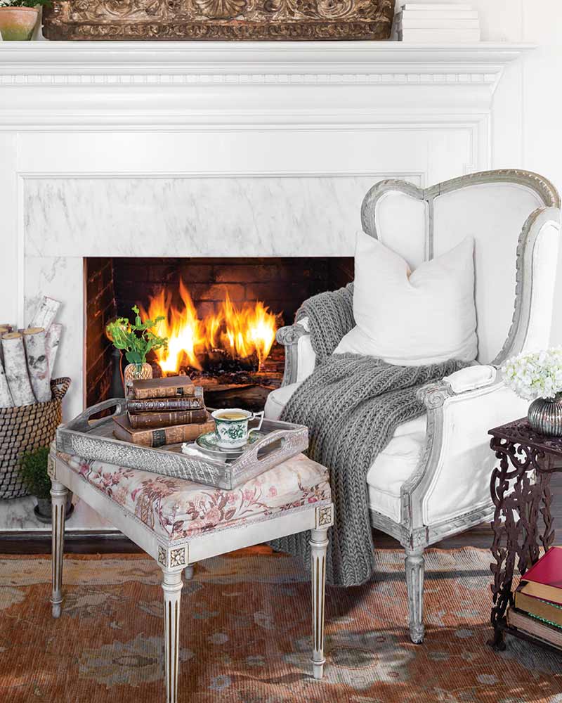A white arm chair nestled near the fire with a cup of tea and blanket.
