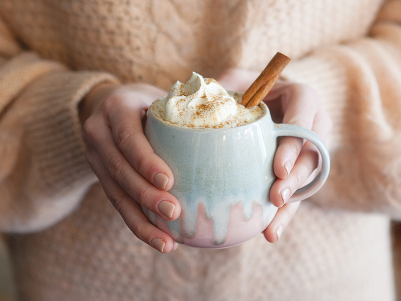 Someone holding a mug of Spiced White Hot Chocolate with a cinnamon stick.