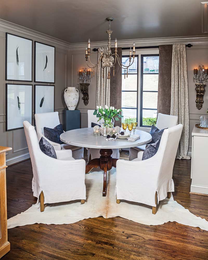 A dining room with a round table, upholstered chairs, and feather watercolor artwork.