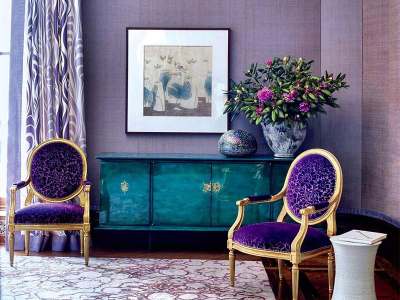 A living room with purple wallpaper, drapes, rug, chairs, and a teal sideboard.