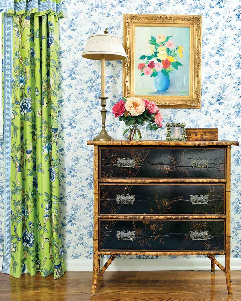 An antique chest in a room with blue patterned wallpaper and blue-and-green drapes.