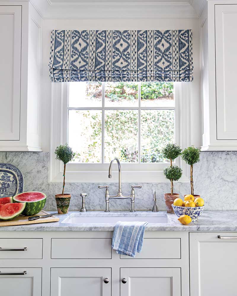 A white kitchen with marble countertops and a blue-and-white ikat roman shade. 