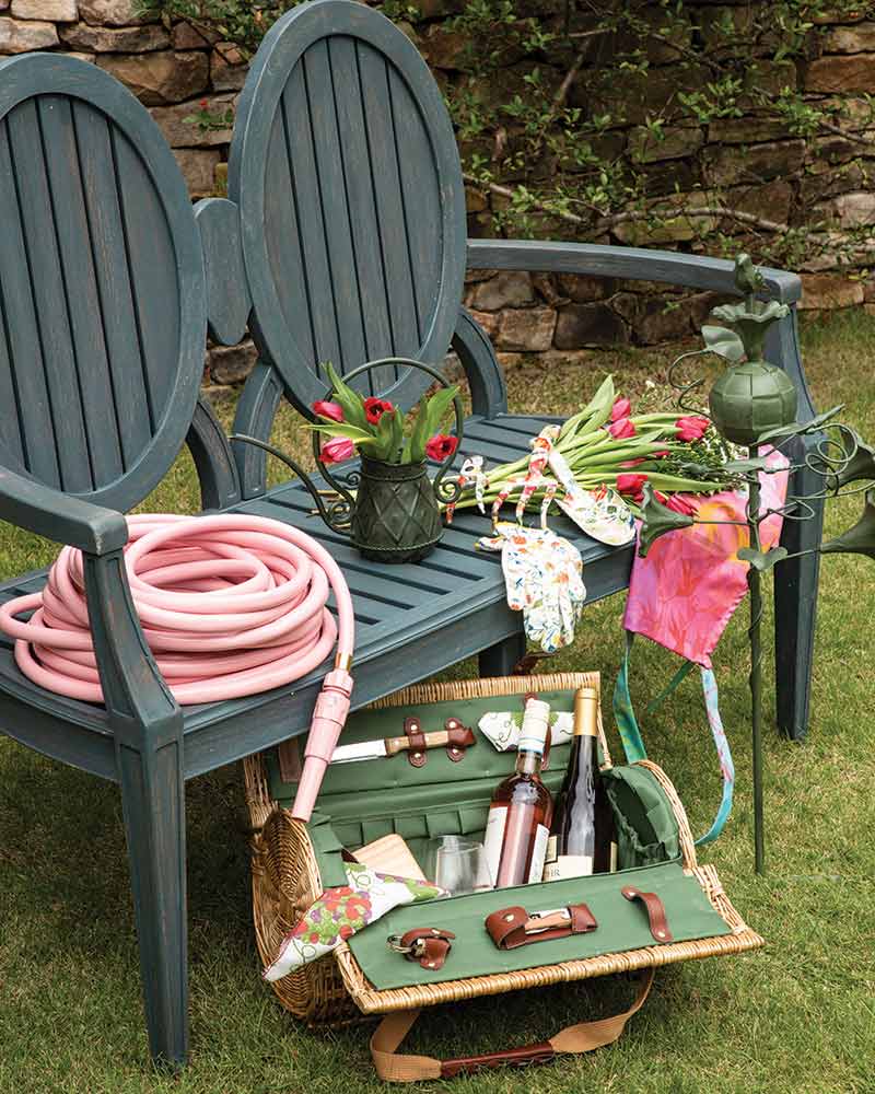 A green bench with a pink gardening hose, rattan picnic basket, and floral gardening tools.