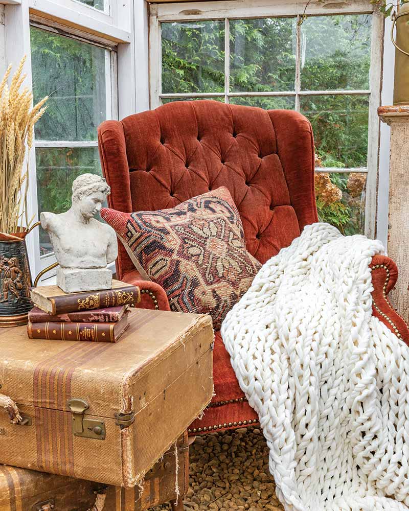 A rust armchair with a whit knit blanket in the corner of a greenhouse.